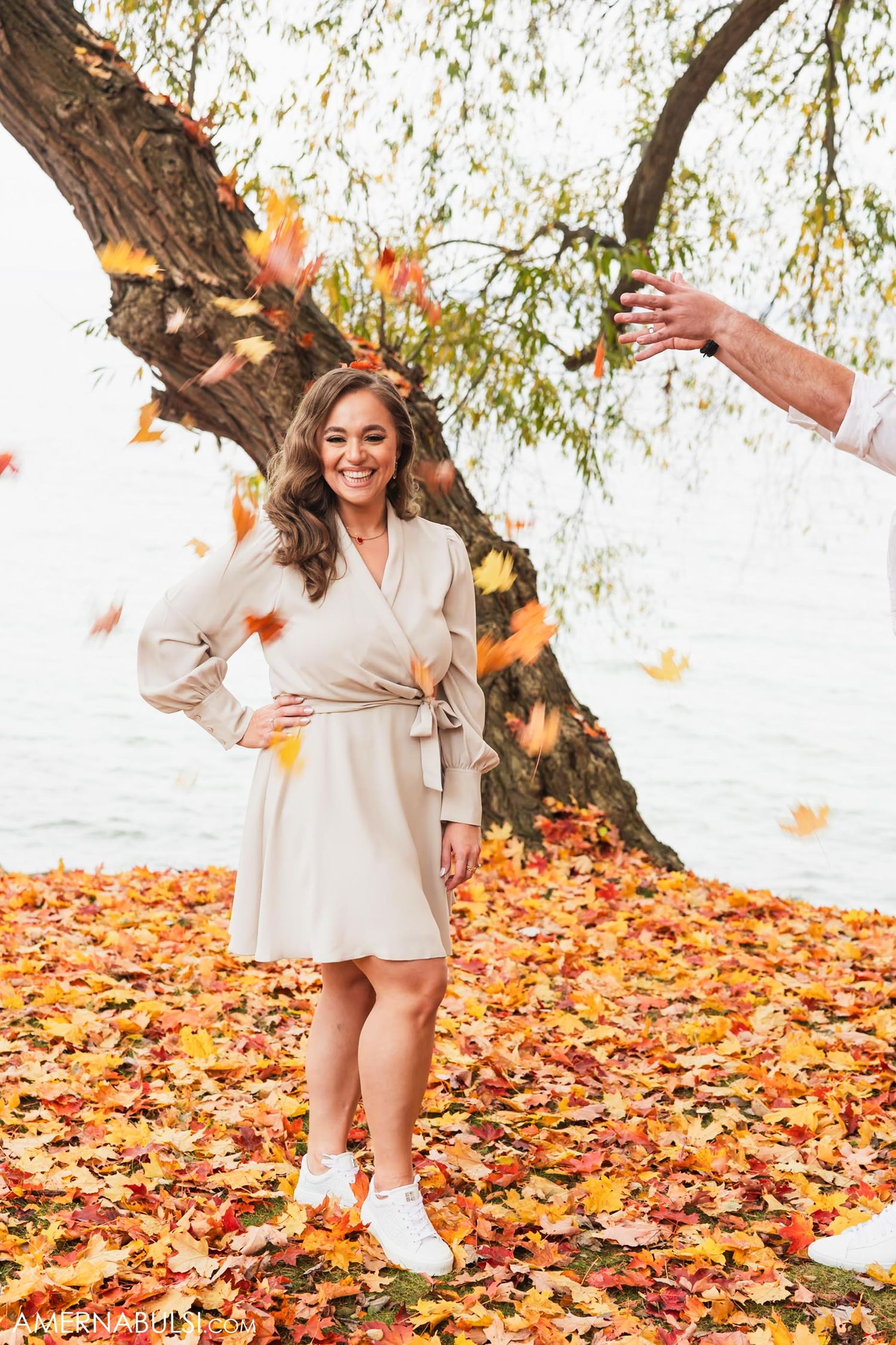 A woman in a cream dress poses in fallen autumn leaves while someone tosses more leaves in the air.