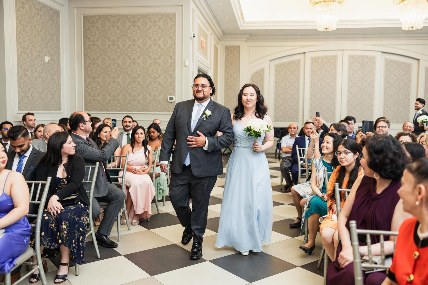 Wedding party members walk down black and white checkered aisle while guests watch in elegant ballroom.