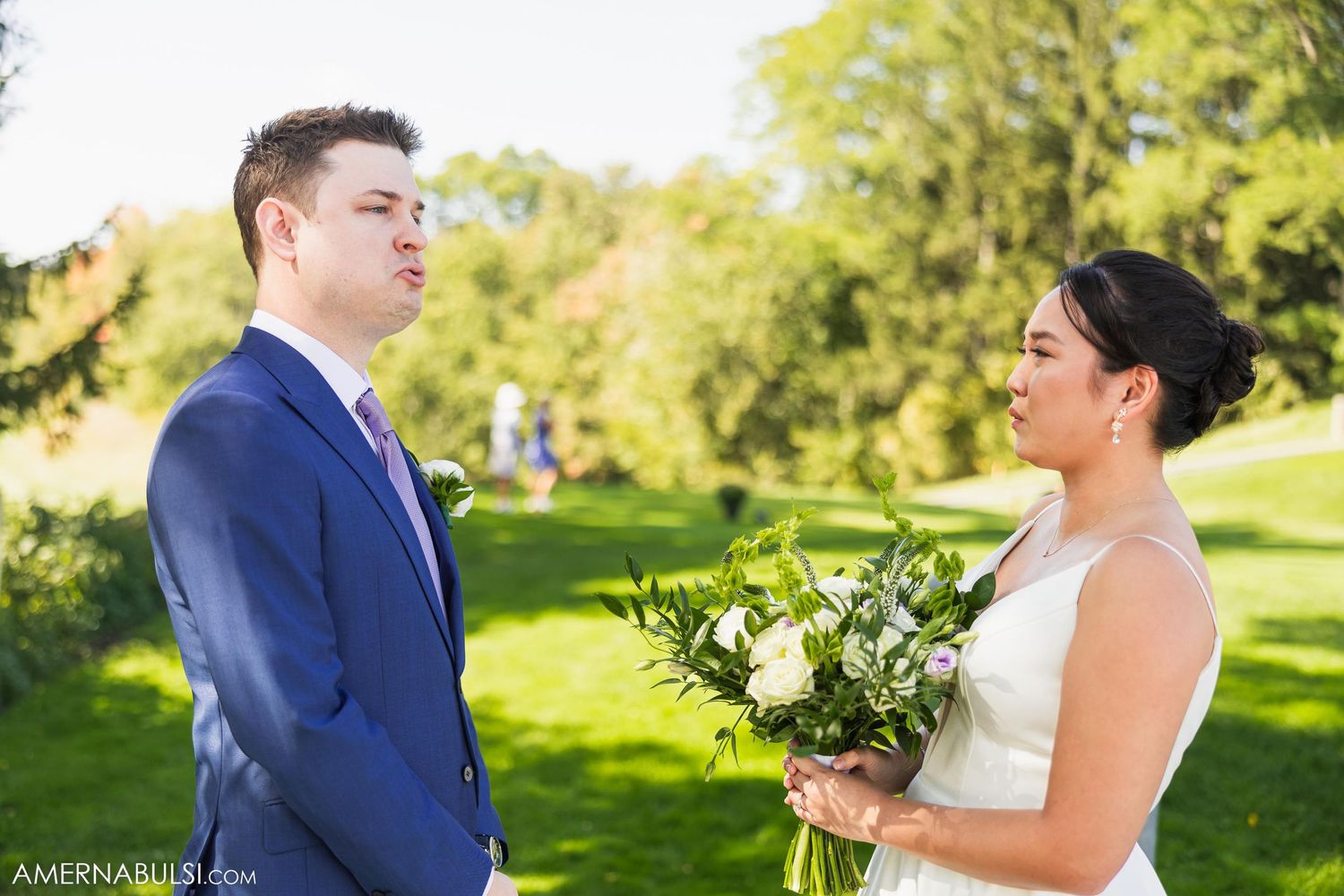 Couple exchanges glances during outdoor wedding ceremony surrounded by green trees.