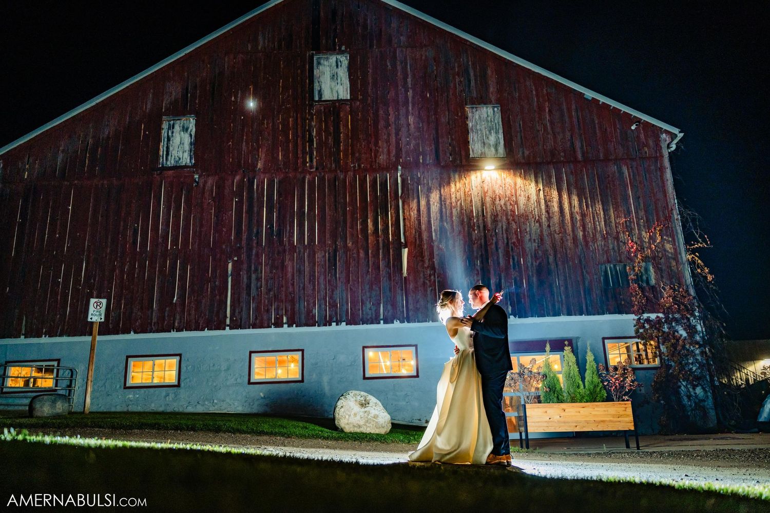 A nighttime wedding photo in front of a rustic red barn with glowing windows.