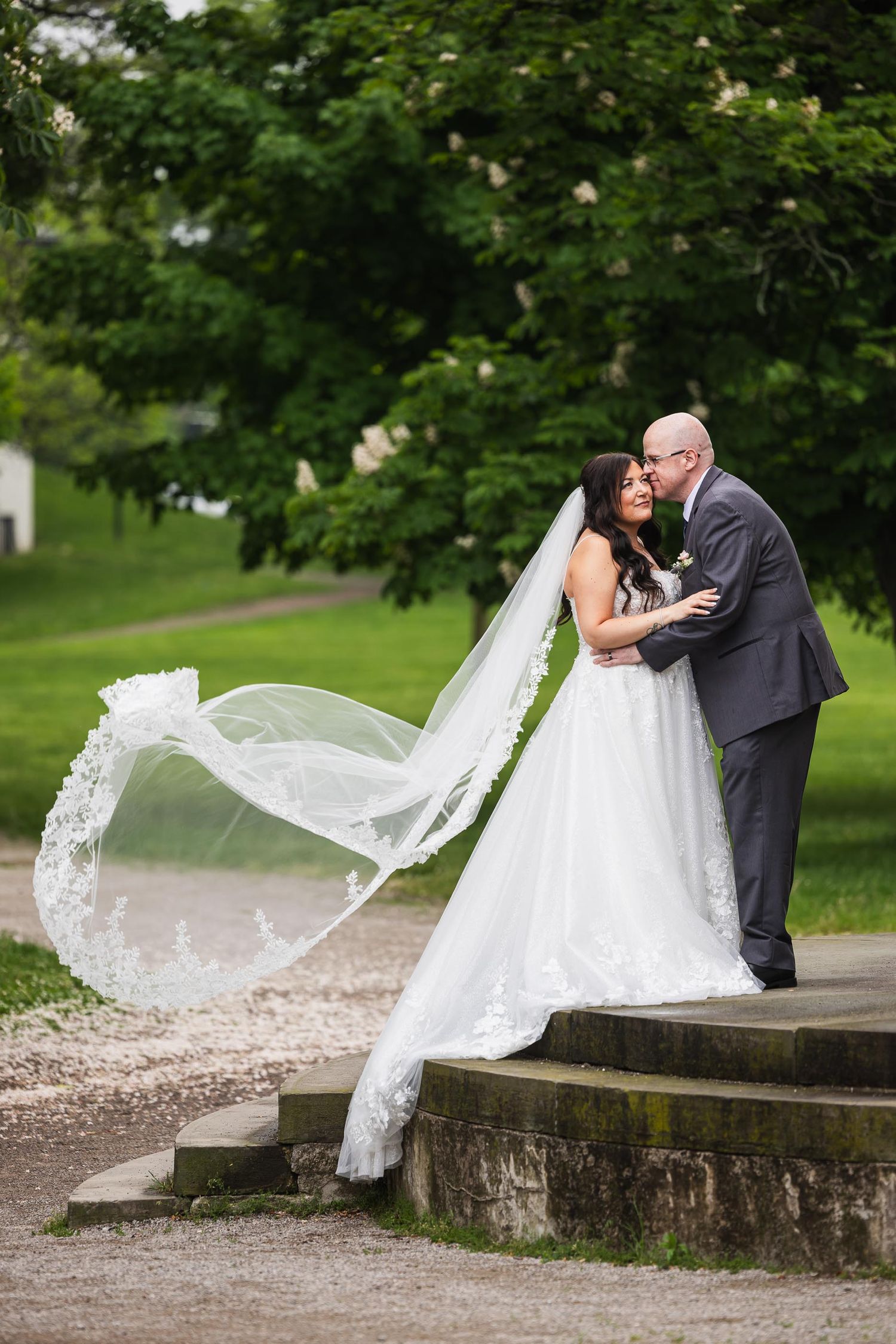 Dramatic wedding photo with bride's veil flowing in the wind as couple embraces on stone steps.