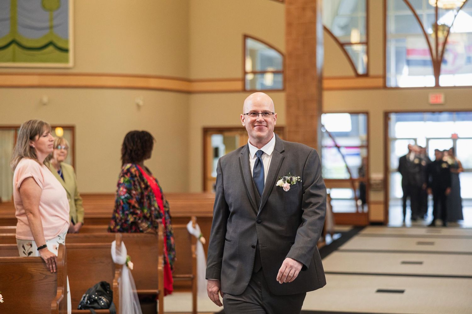 A person in a grey suit with boutonniere stands in a church during a wedding ceremony.