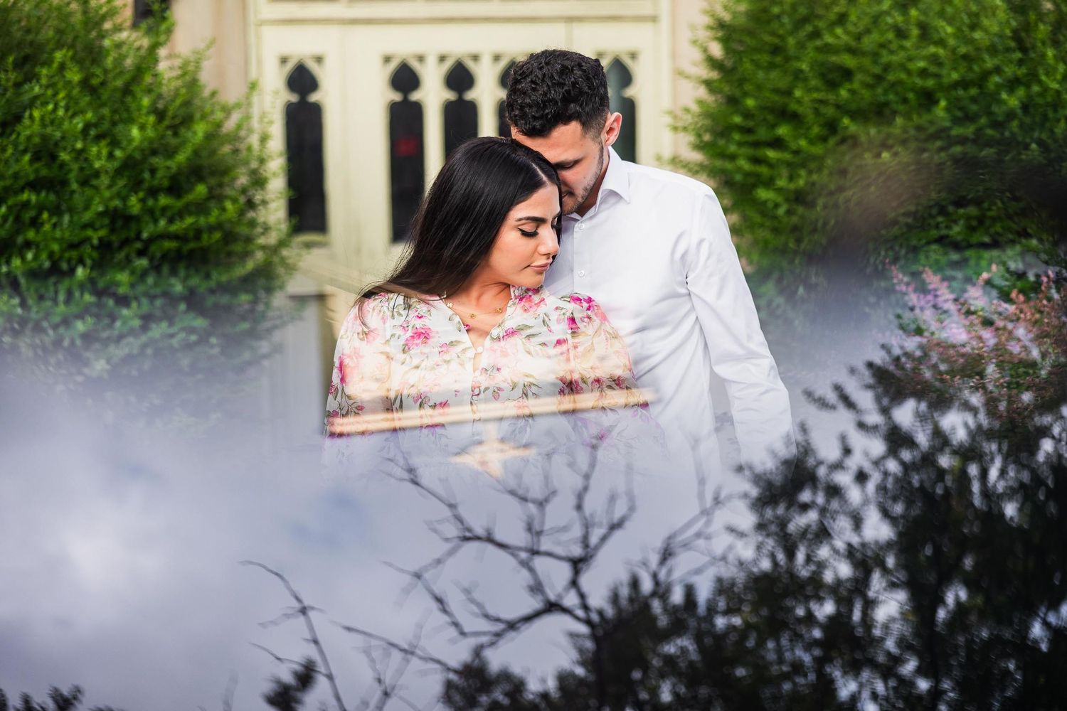 A romantic couple embraces in front of Gothic architecture with misty floral gardens in the background.