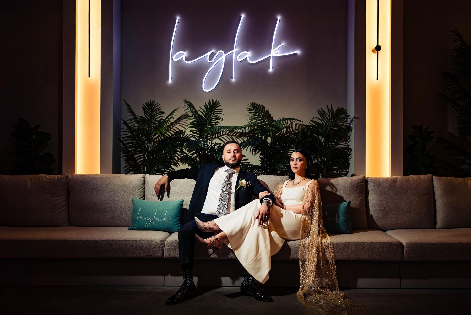 A couple relaxes on a couch beneath an illuminated neon 'light' sign during an elegant evening celebration.