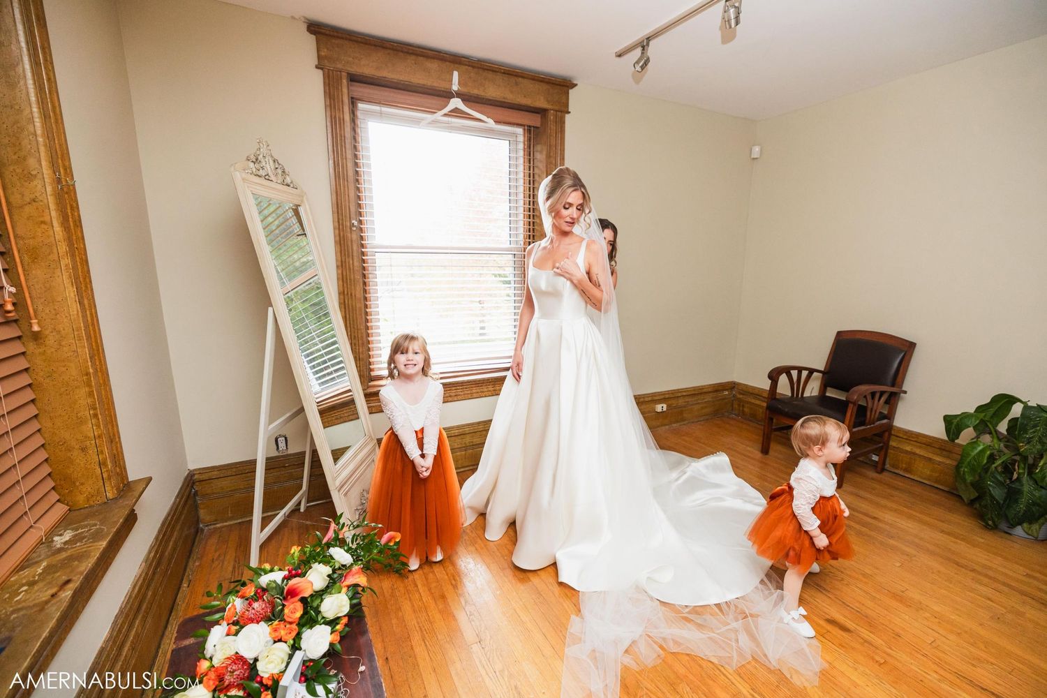 Bride in elegant white gown stands with two small attendants in orange dresses near a vintage mirror.