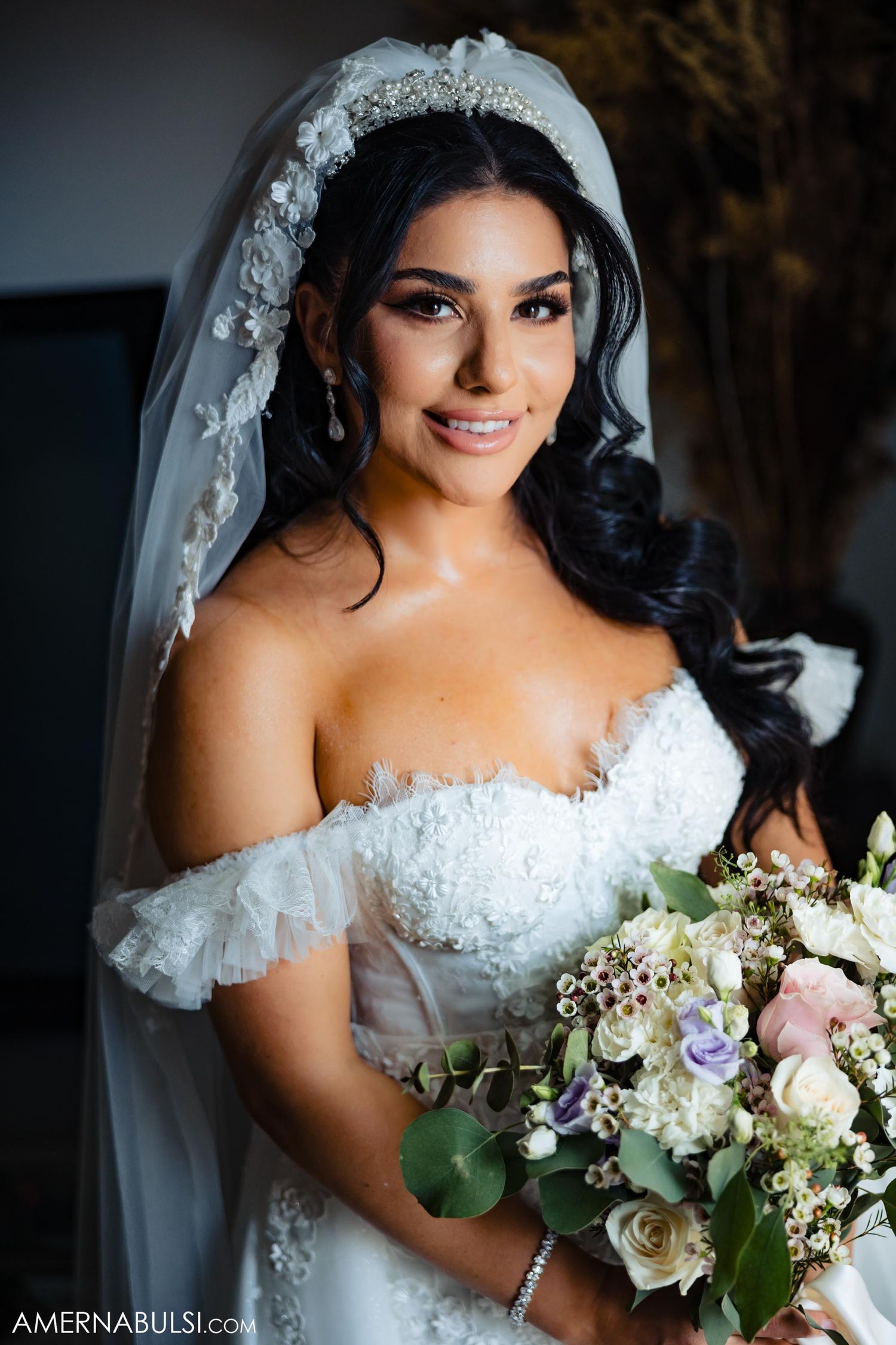 Bride wearing lace off-shoulder gown and veil holds elegant bouquet while posing indoors.