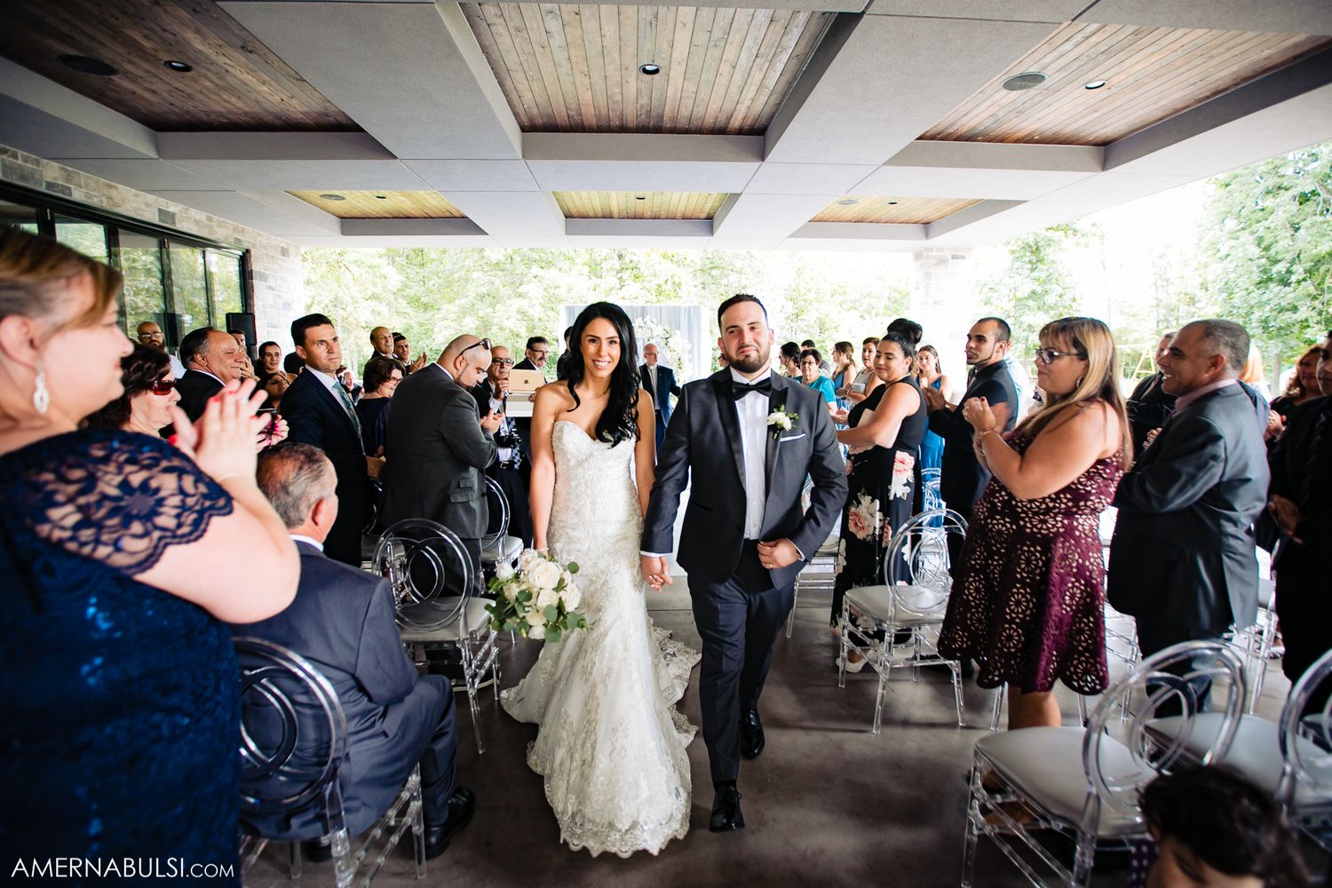 Wedding guests enjoy an outdoor reception under a covered patio area with natural greenery in the background.