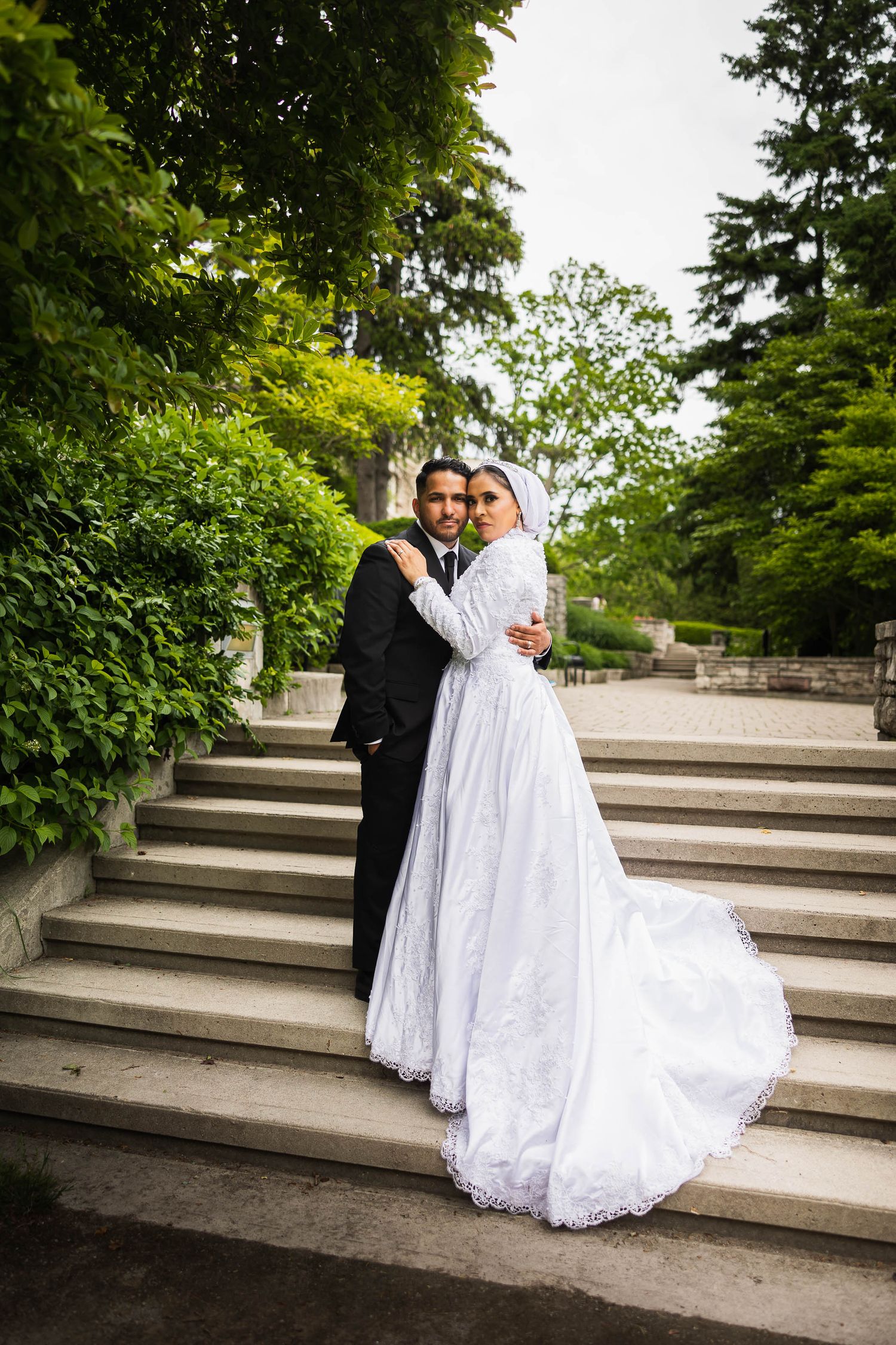 A wedding couple poses elegantly on stone steps surrounded by lush greenery.