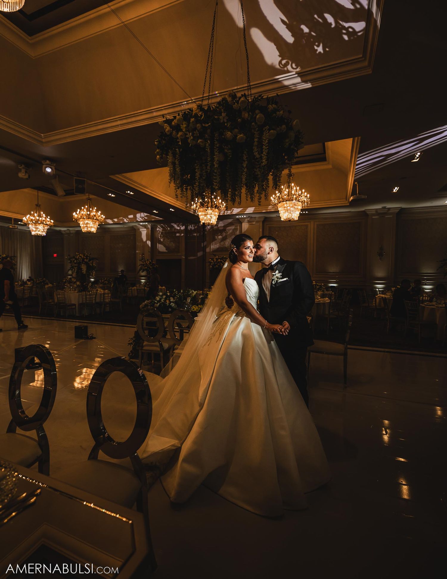 Romantic wedding couple pose under crystal chandeliers in an elegantly lit ballroom.