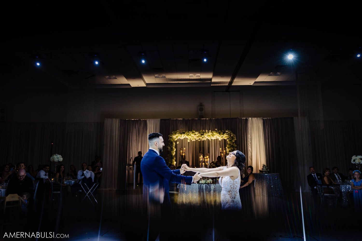 Dramatic spotlight illuminates a couple's first dance in a dark reception hall with guests watching.