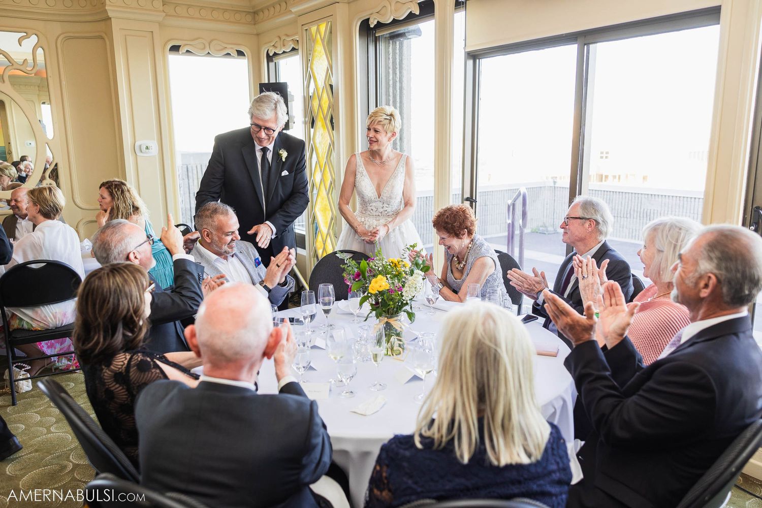 Wedding reception scene with guests applauding at round tables decorated with centerpieces.
