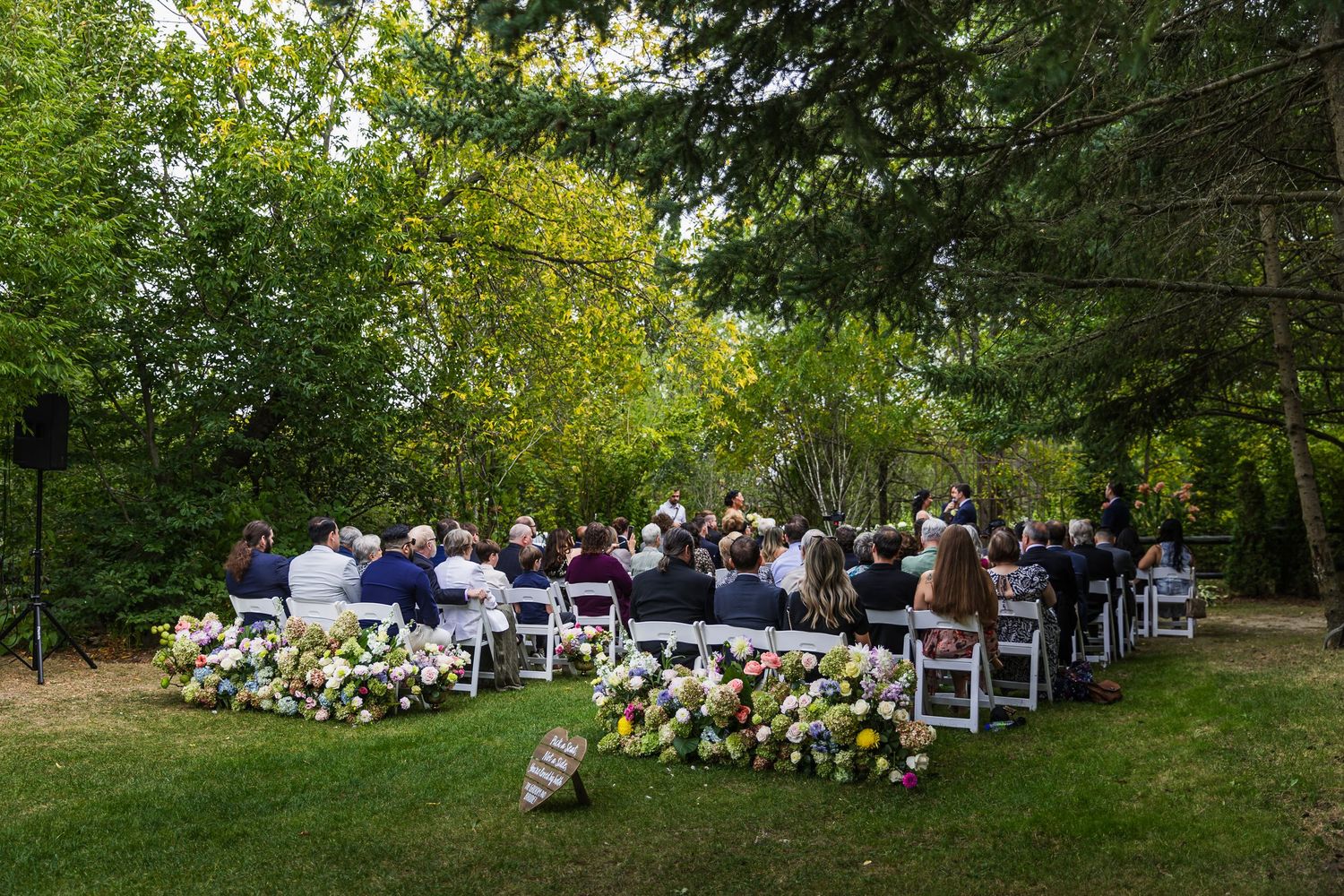 Outdoor wedding ceremony with guests seated in white chairs surrounded by lush green trees and floral arrangements.