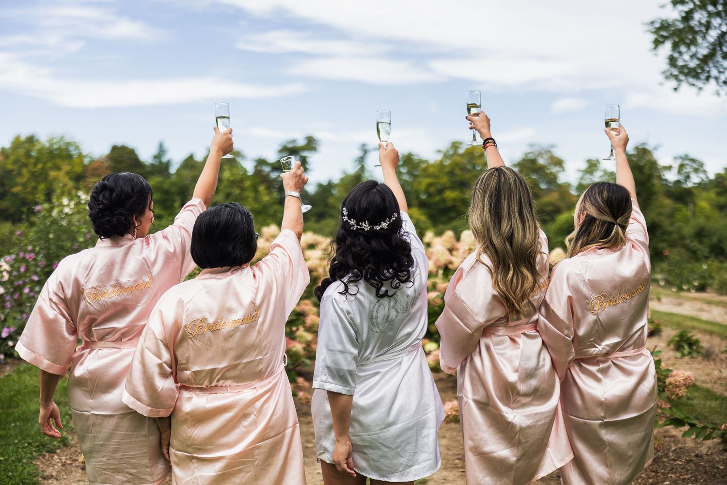 Bride and bridesmaids in satin robes raising champagne glasses in a celebratory toast outdoors.