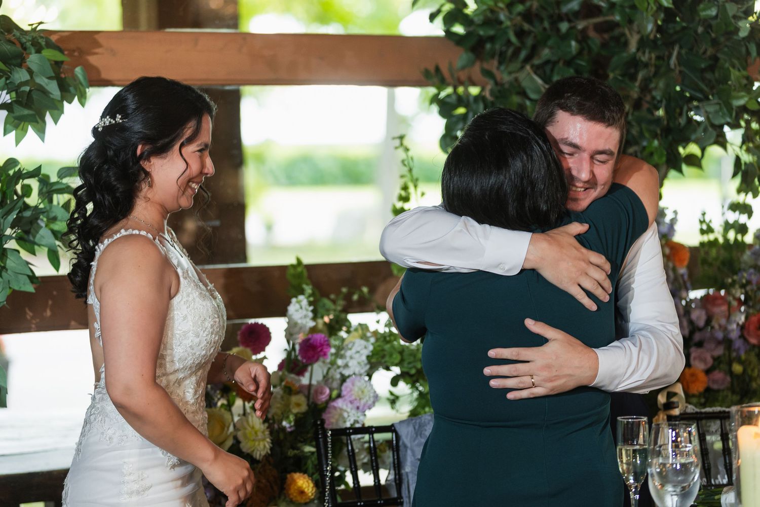 Bride smiles as groom hugs a guest at an elegant wedding reception with floral centerpieces.