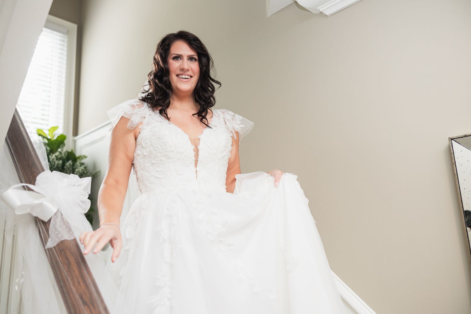 Smiling bride in lace ball gown holding dress while descending staircase before wedding.