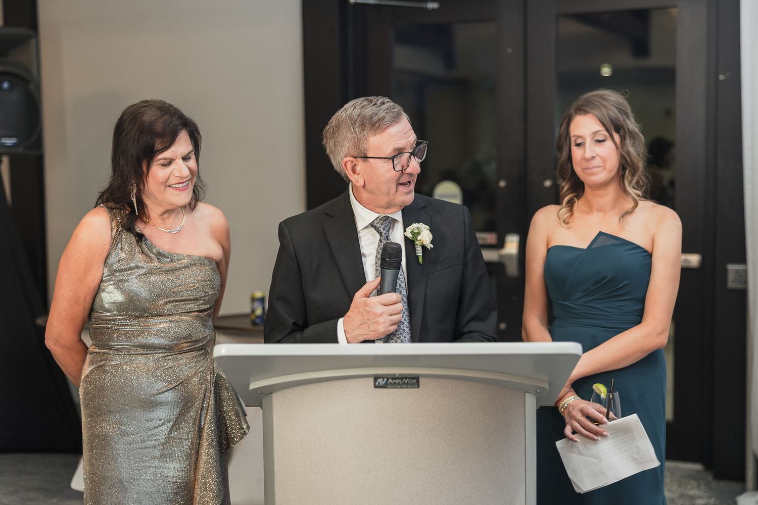 Man giving speech at podium flanked by two women in formal attire at an elegant wedding reception.