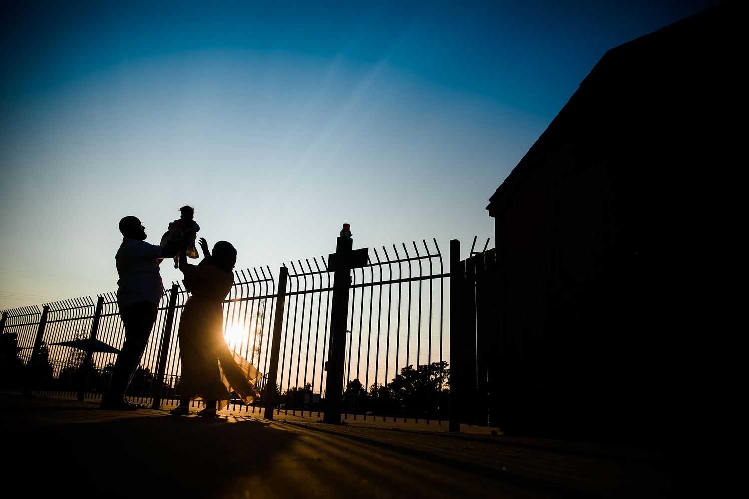 Silhouettes of people against sunset sky with iron fence railing creating dramatic backlit scene.