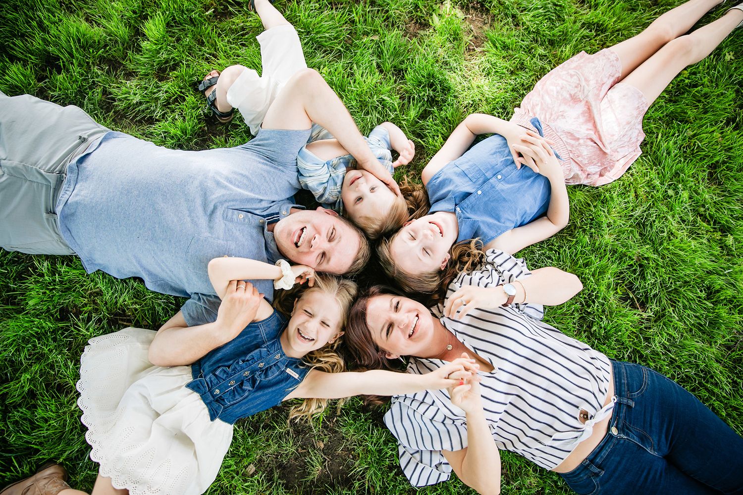A family lies together on green grass in a playful moment during a summer photoshoot.