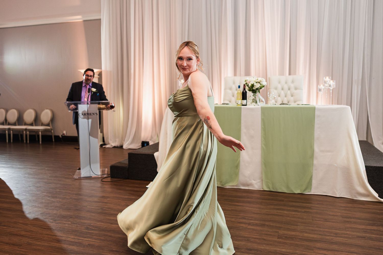 Bridesmaid in sage green gown twirls on dance floor while officiant speaks at wedding reception.