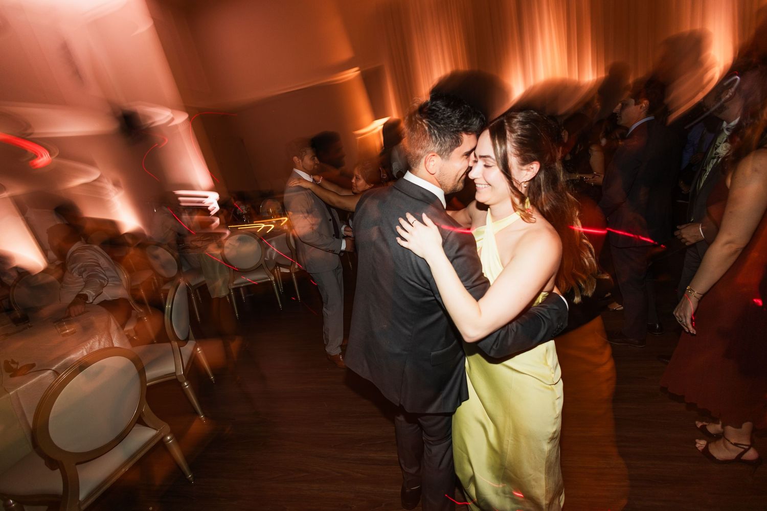 Couple shares a romantic dance at a wedding reception with warm amber lighting and guests in the background.