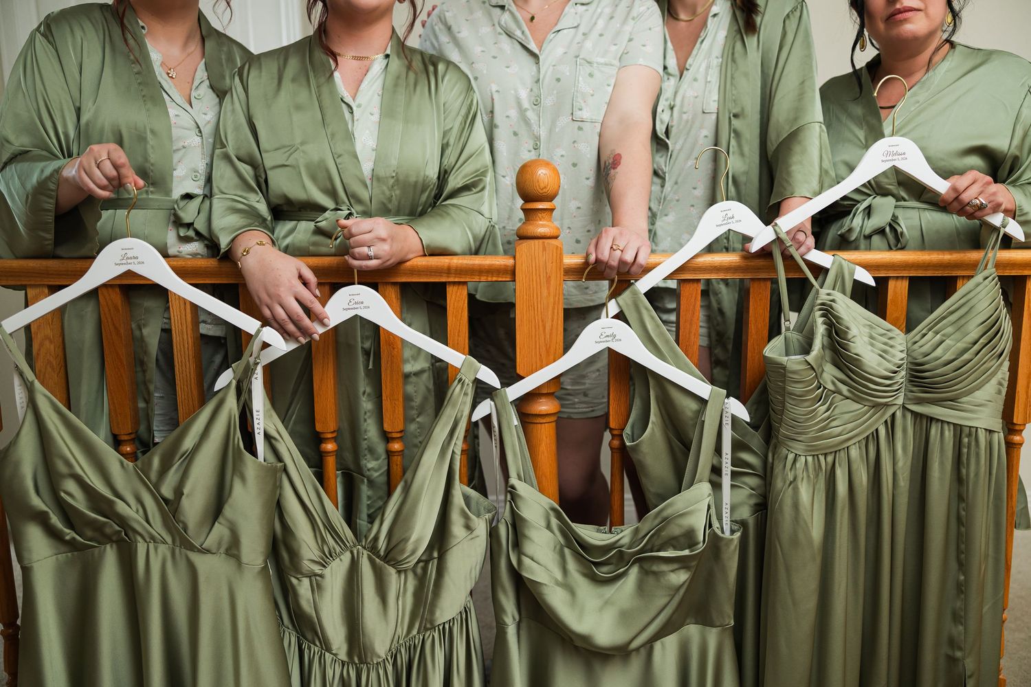 Bridesmaids in sage green robes hold hangers displaying elegant olive green satin bridesmaid dresses before a wedding.