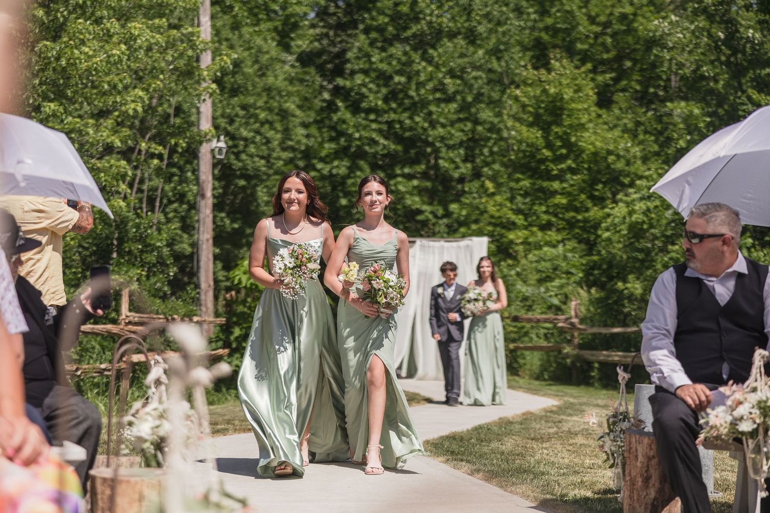 Bridesmaids in sage green dresses walk down outdoor wedding aisle carrying bouquets among lush green trees.