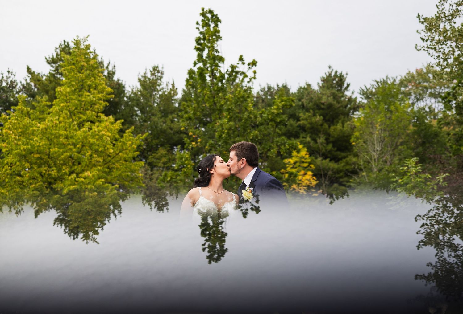 Bride and groom sharing romantic kiss outdoors surrounded by lush green trees with misty reflection effect.