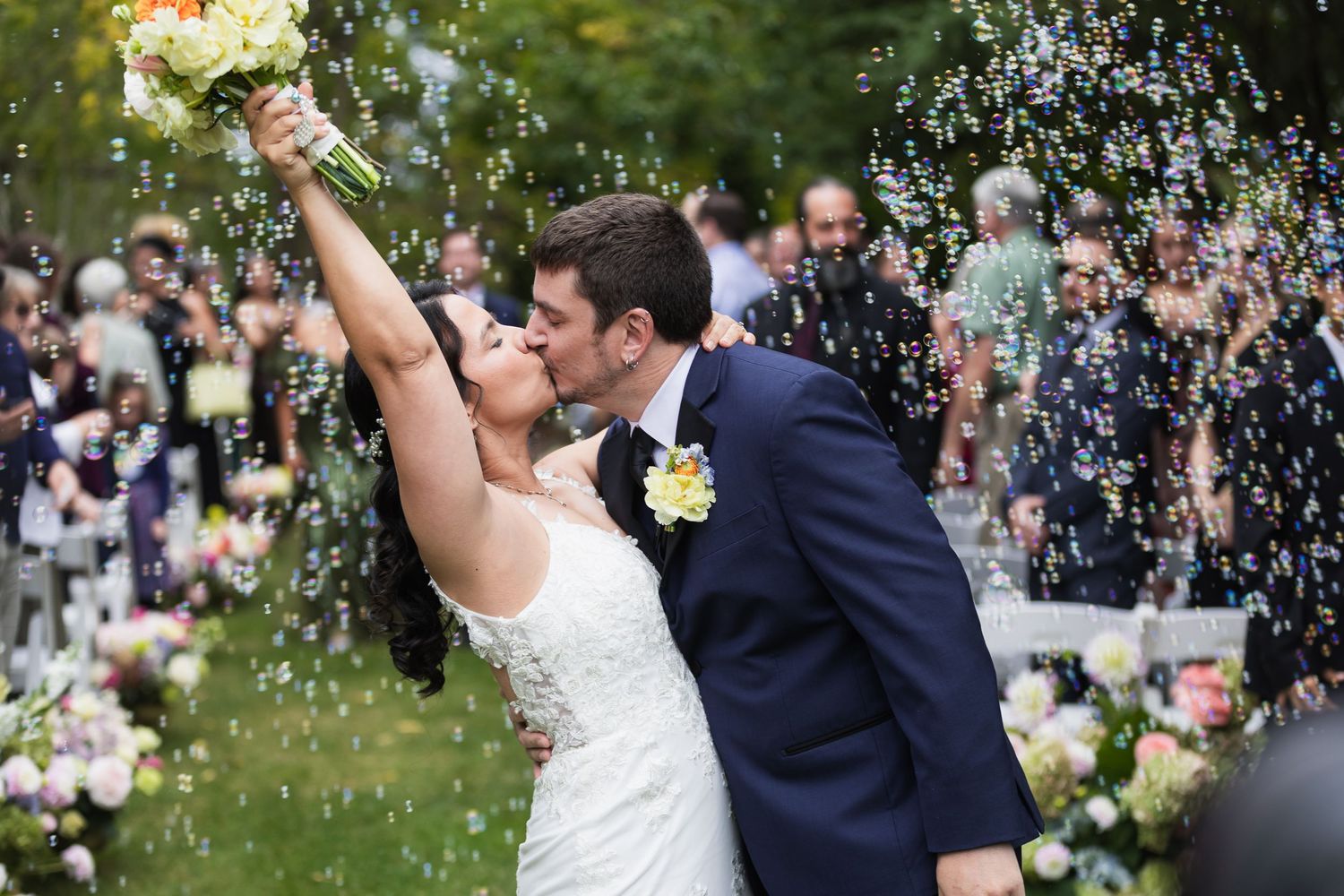 Bride and groom share a kiss surrounded by bubbles at outdoor wedding ceremony, bride holding yellow bouquet.