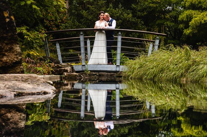 Wedding couple poses on garden bridge overlooking tranquil pond with lush greenery.