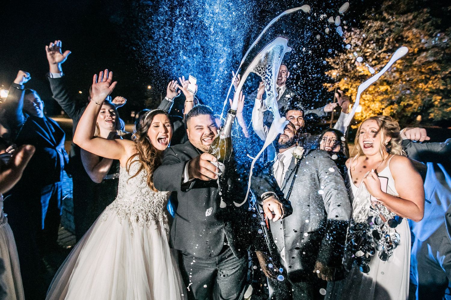 Celebratory wedding party moment with champagne spray creating dynamic splash effect at night.