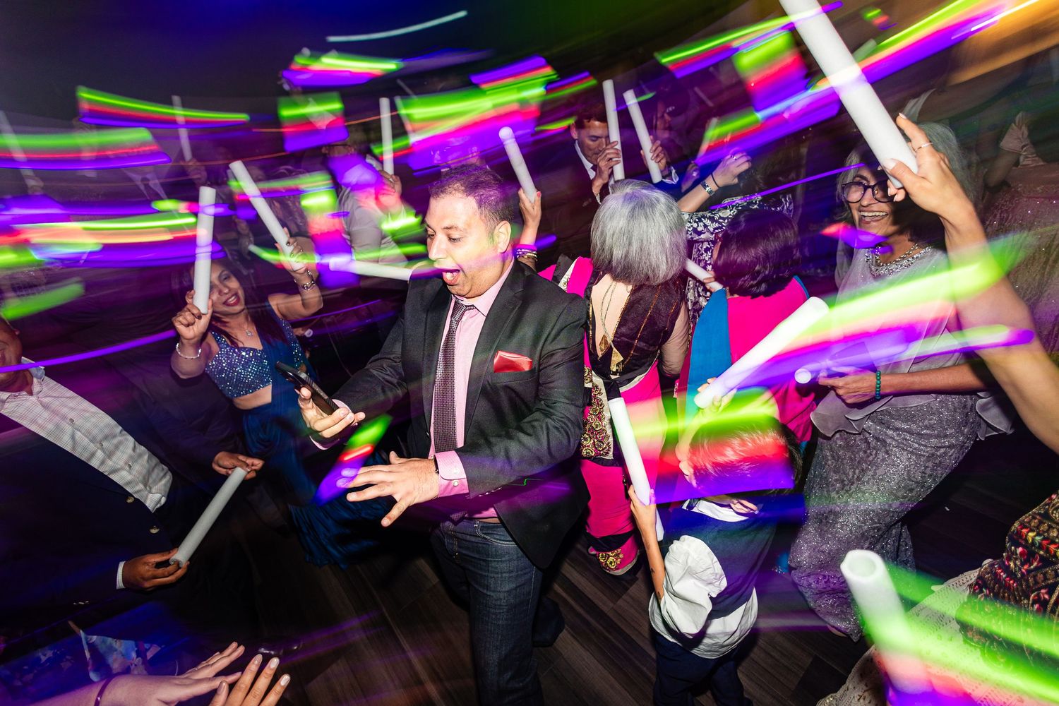 Nightclub crowd dancing with colorful LED light sticks creating vibrant streaks of purple and pink in the dark.
