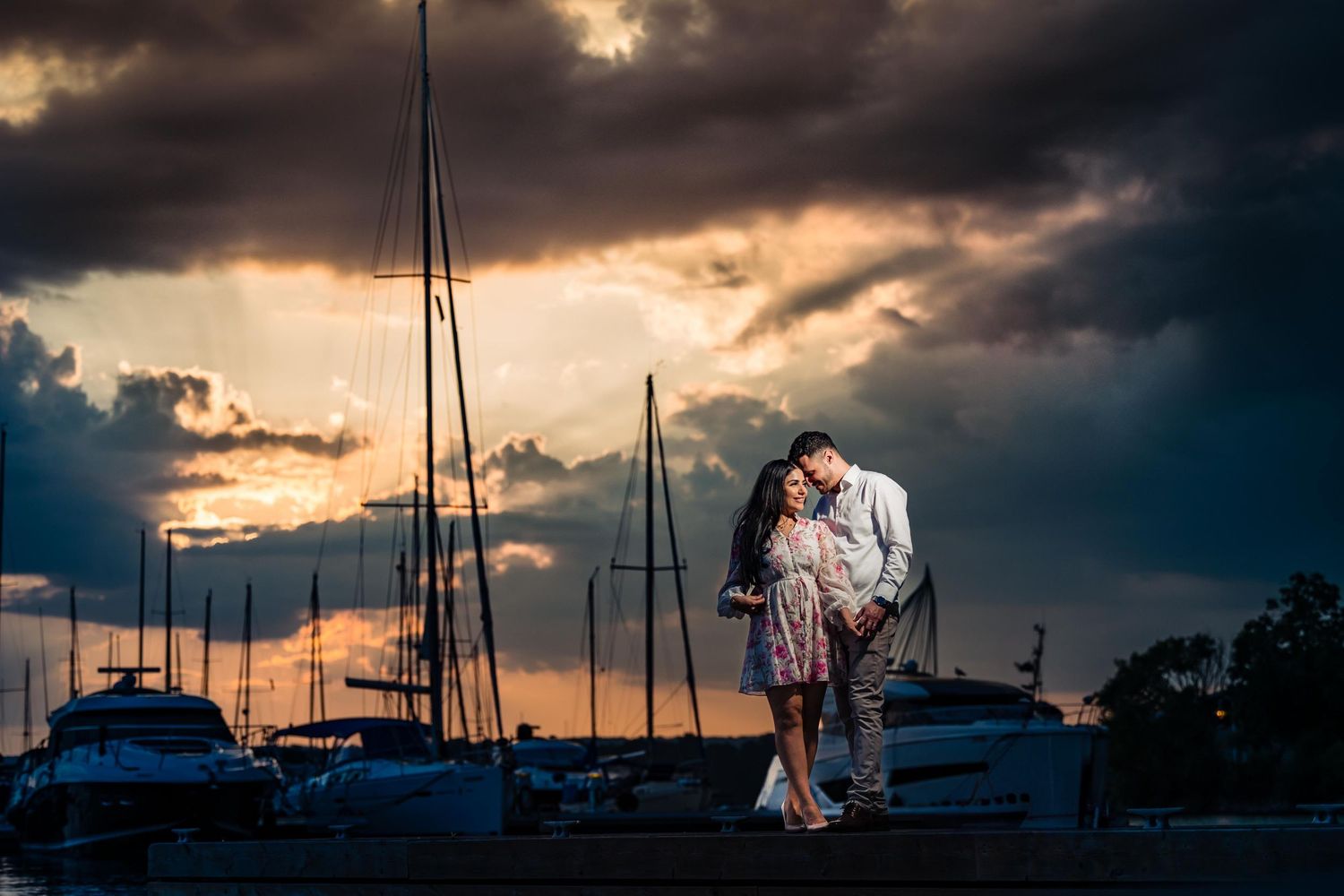A romantic couple embraces at a marina during sunset with dramatic storm clouds and sailboat masts silhouetted in background.
