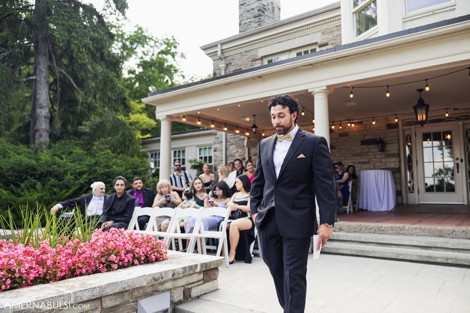 A person in formal black suit walks along a garden path in front of an elegant wedding venue with guests seated.