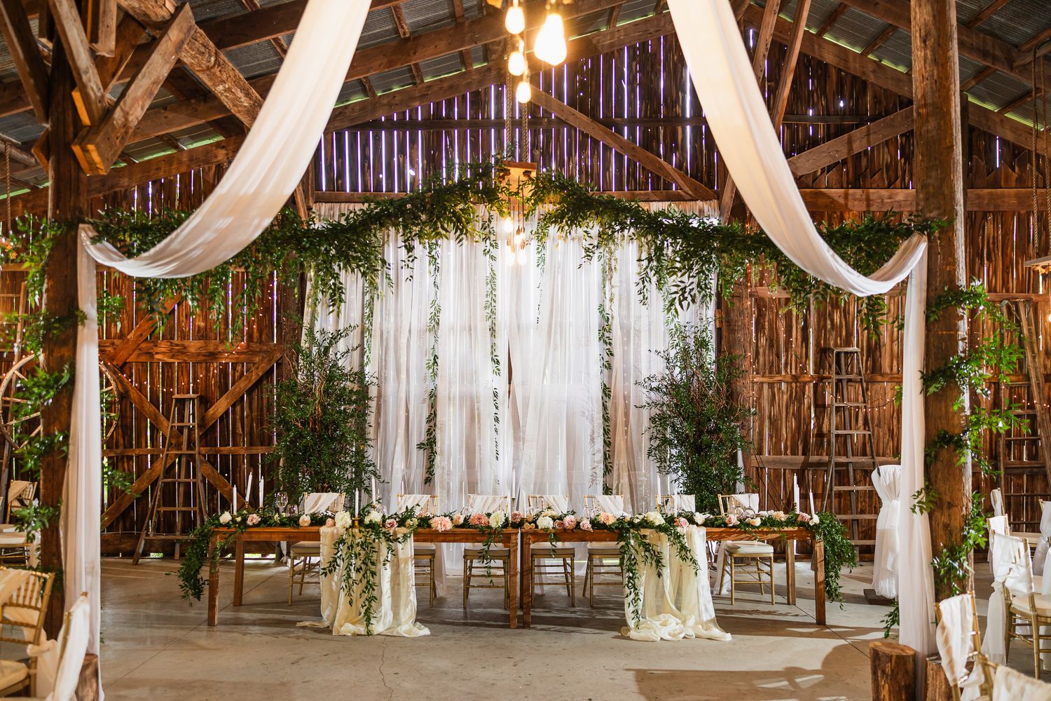 White fabric draping and greenery decorate the ceiling of a rustic wooden barn wedding venue with natural lighting.