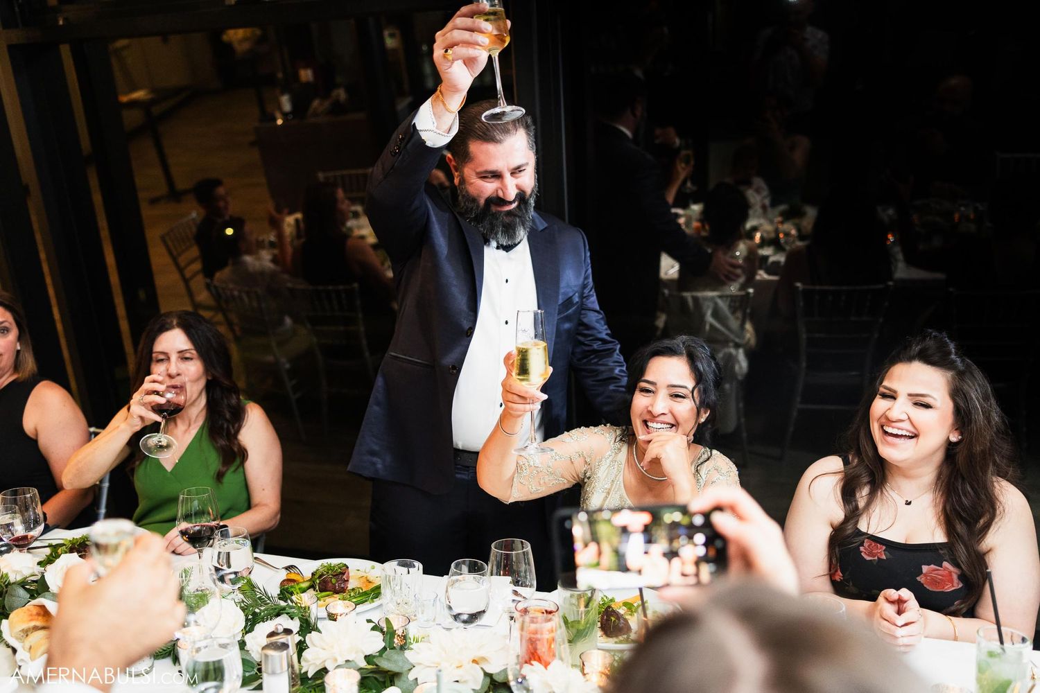 Groom making celebratory toast while standing at wedding reception dinner table.