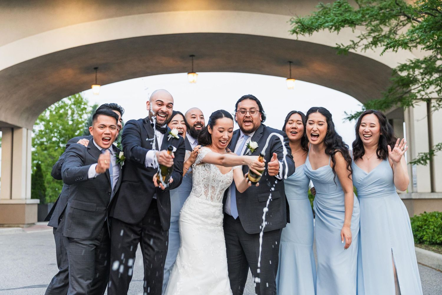 Wedding party poses playfully with champagne bottles under an outdoor arch.