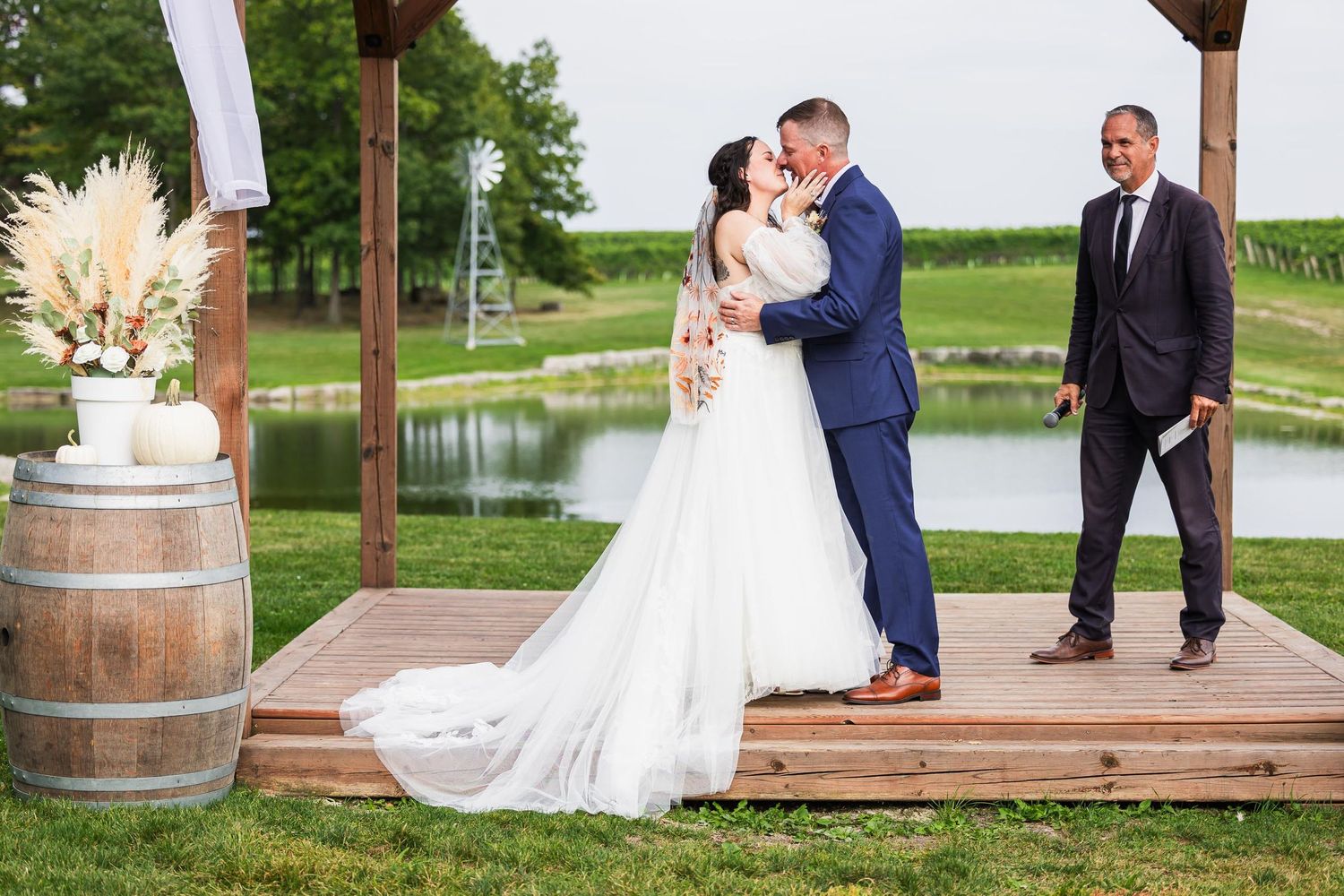 A romantic wedding ceremony takes place on a wooden deck by a pond with a rustic wine barrel and pampas grass decor.