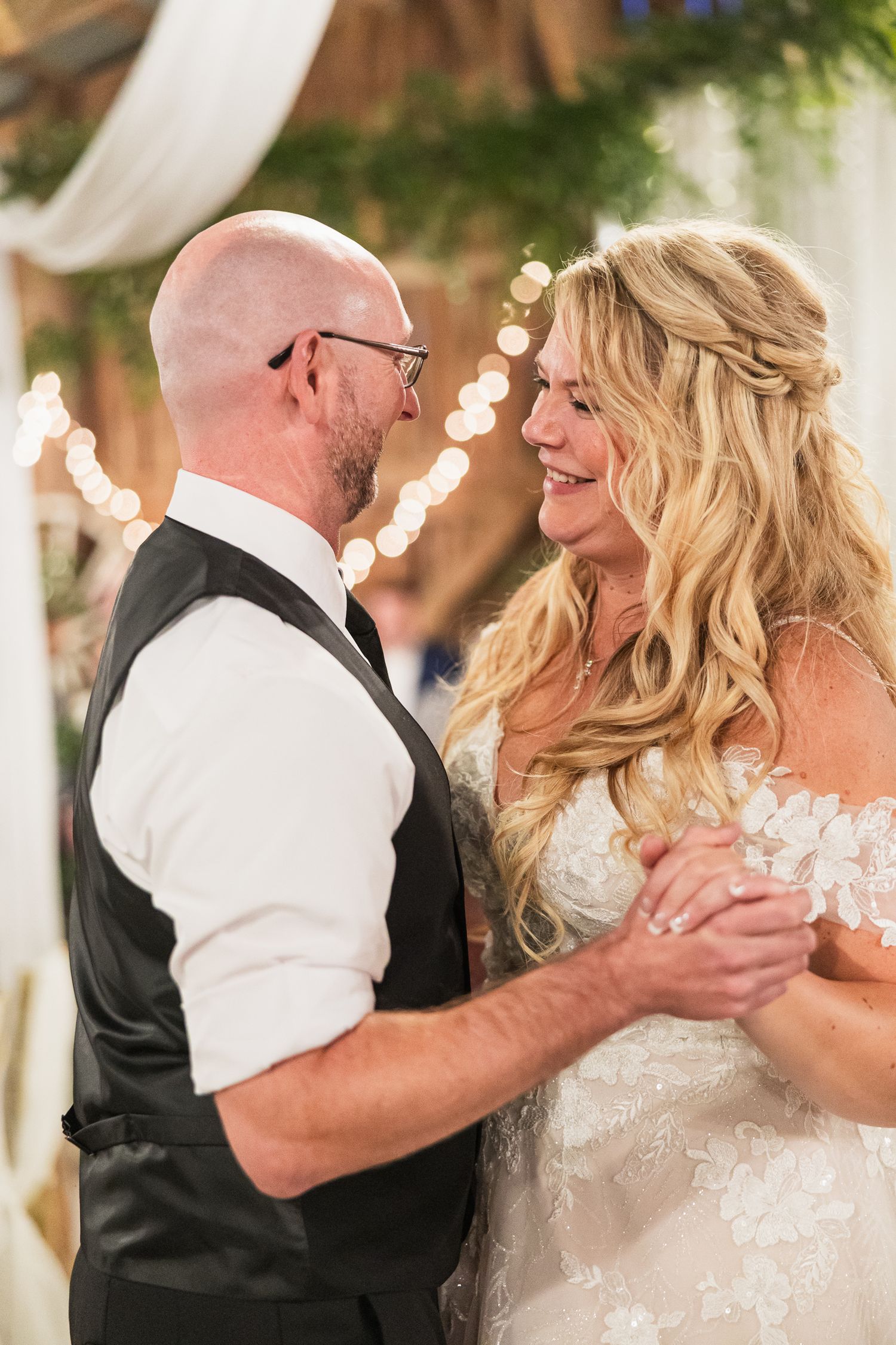 A couple shares a joyful moment during their first dance at a wedding reception with string lights in the background.