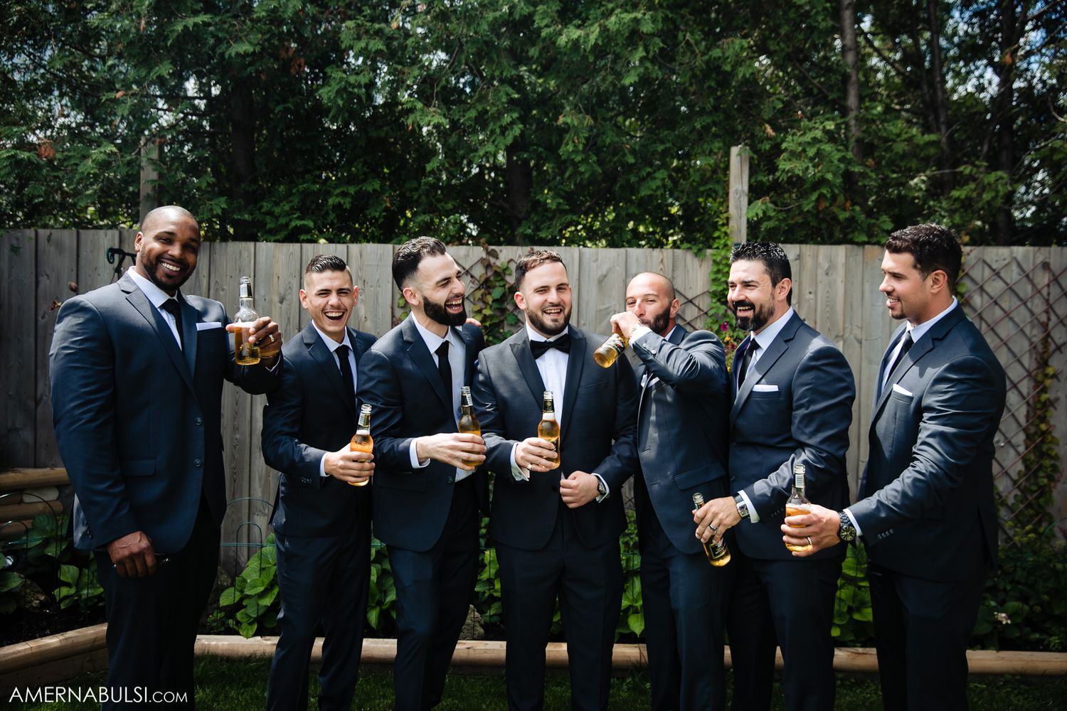 A group in formal navy suits raise beer glasses in celebration during an outdoor wedding reception.