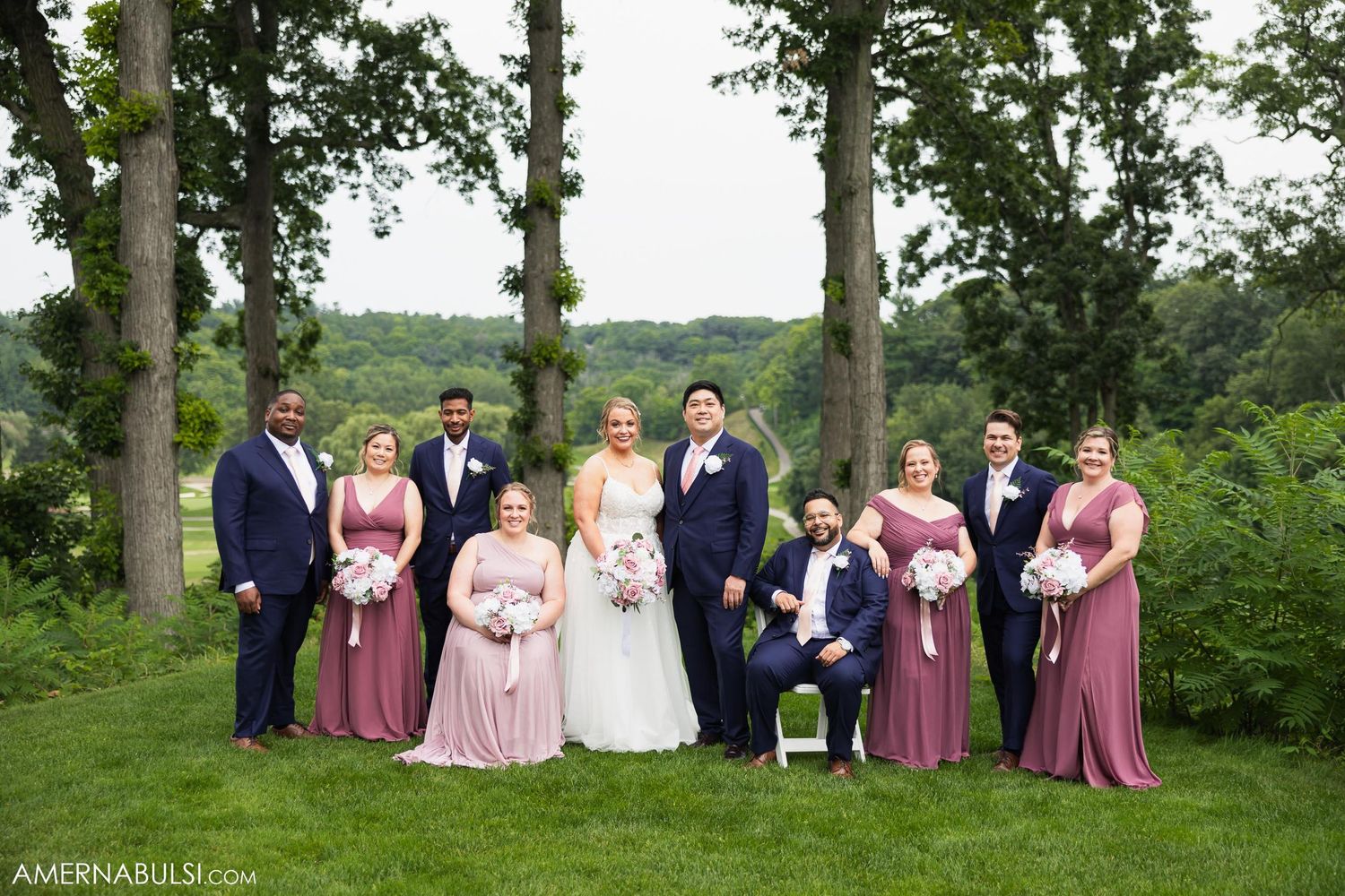 Wedding party poses outdoors with bridesmaids in mauve dresses and groomsmen in navy suits among tall trees.