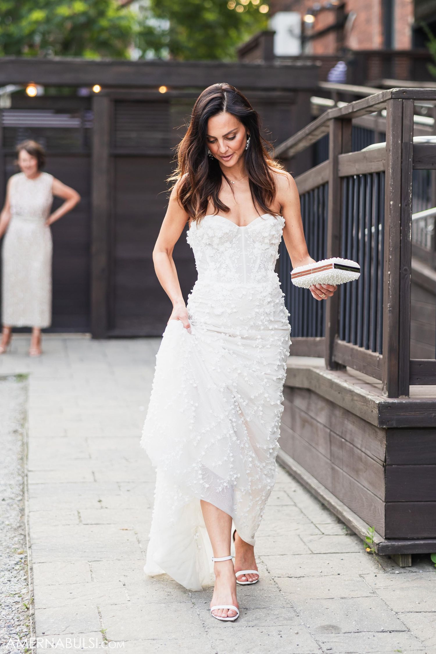 A bride in a white lace dress walks down a staircase while holding a plate in an outdoor setting.