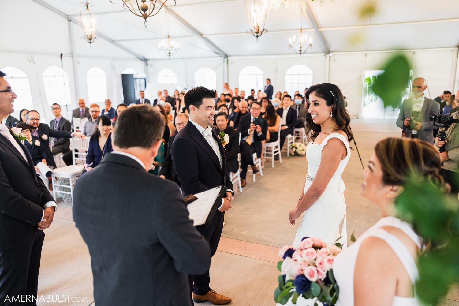 Indoor wedding ceremony with guests seated in white chairs and elegant chandelier lighting.