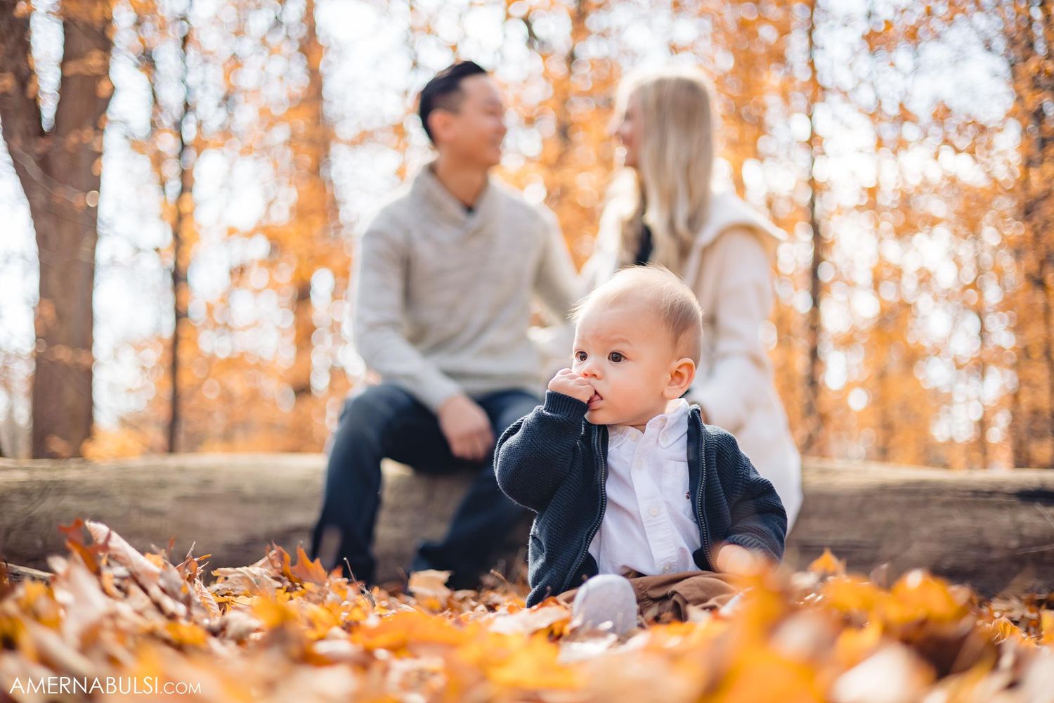 Baby crawls in fallen leaves while parents sit on log in autumn forest.