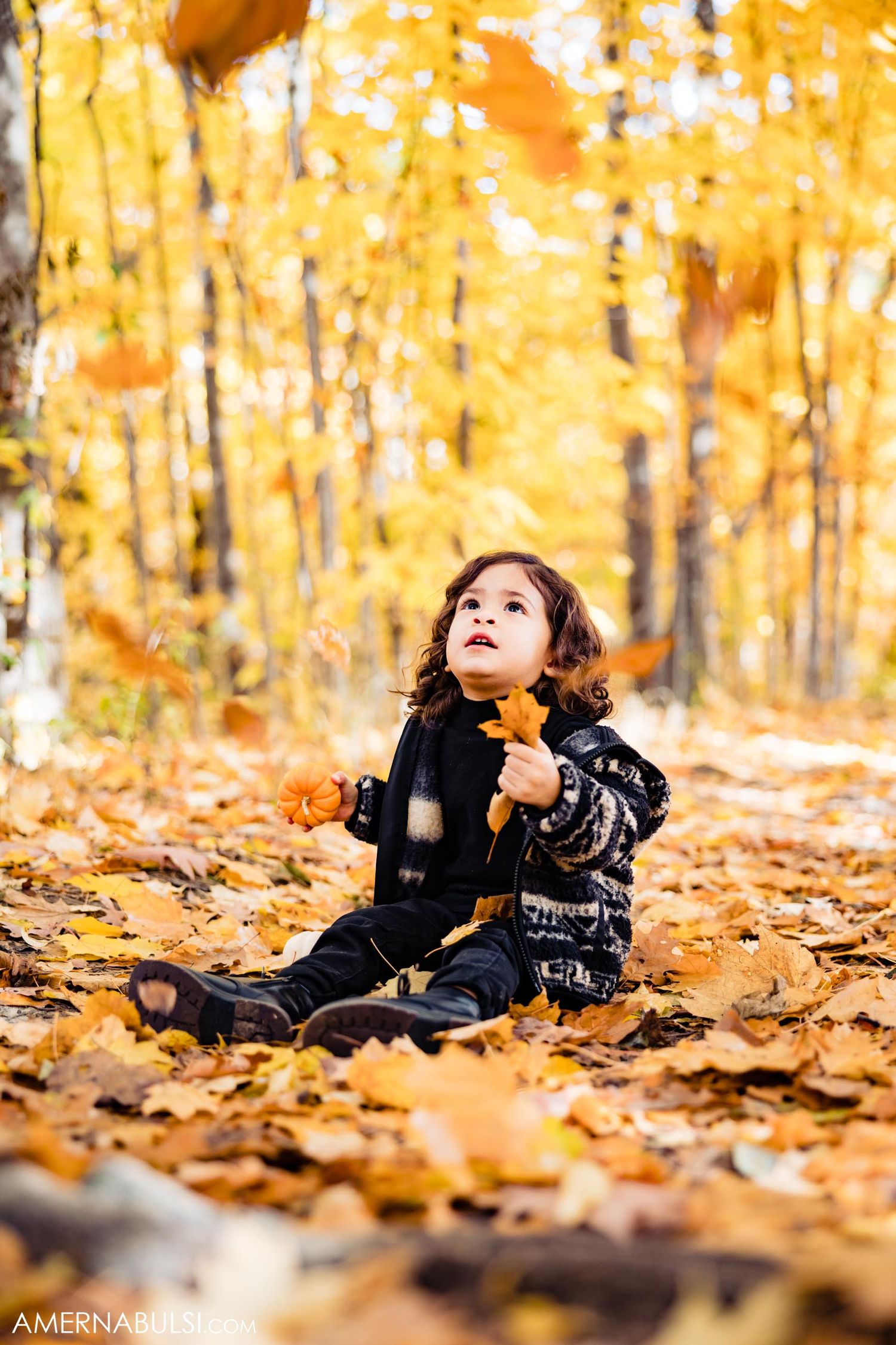 A child plays with fallen leaves during an autumn photoshoot in a golden forest setting.