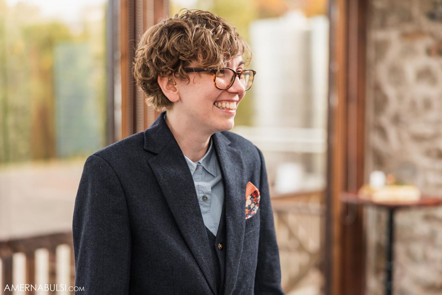 Person in formal attire smiling while wearing black suit jacket and glasses.