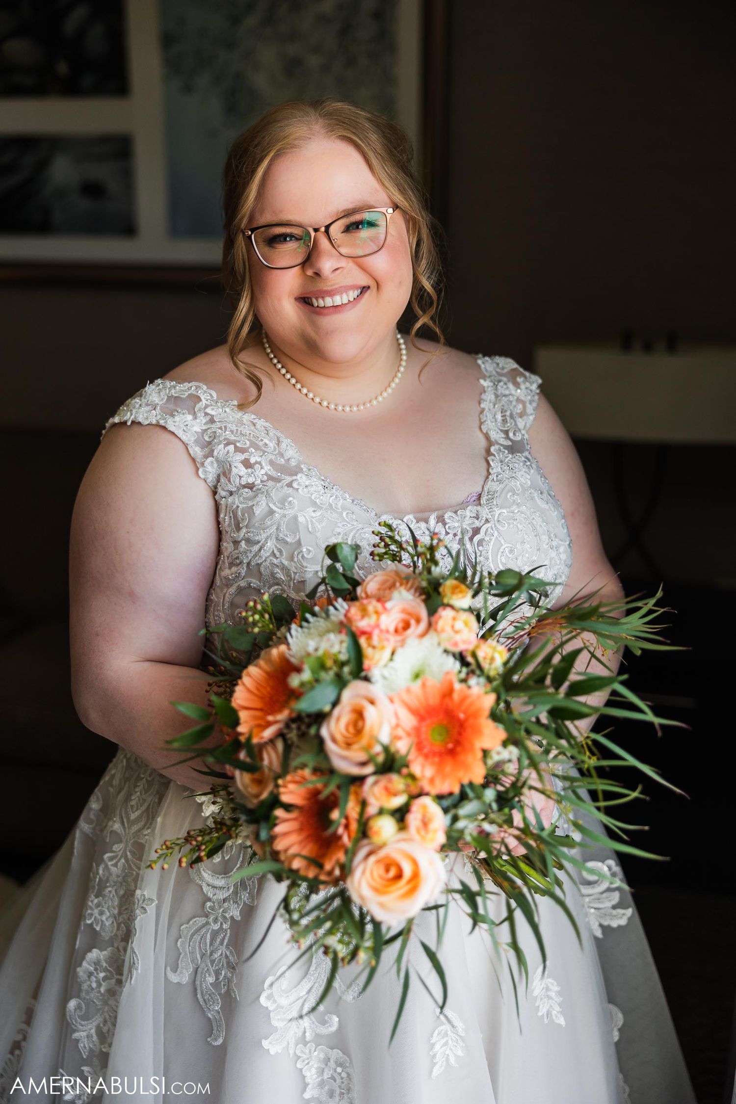 Bride in lace detailed dress holds vibrant peach and orange bouquet indoors.
