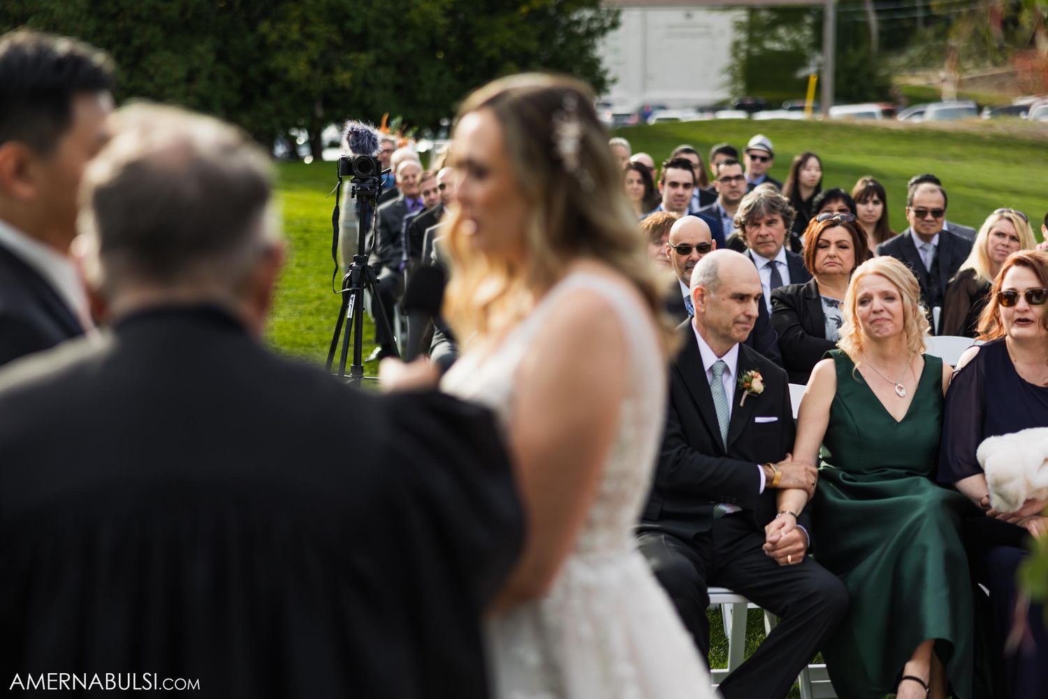 Guests watching an outdoor wedding ceremony from white chairs arranged on a grassy lawn.