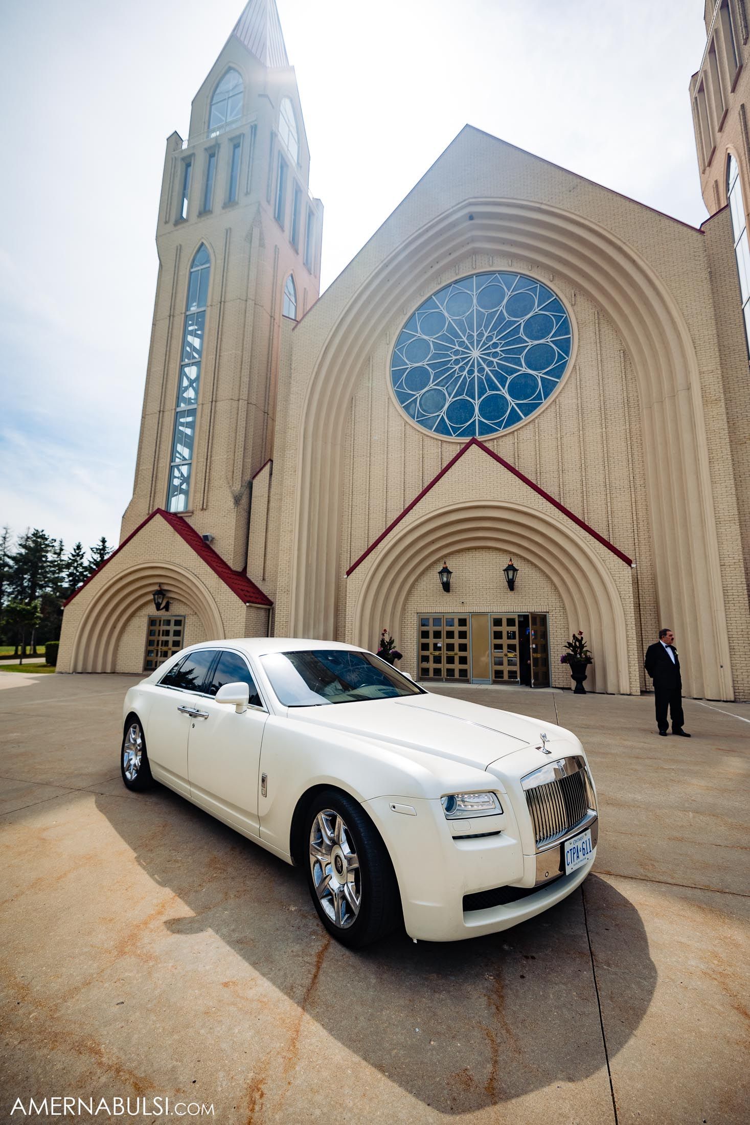 A white Rolls Royce Ghost parked in front of a modern Gothic church with a blue rose window.
