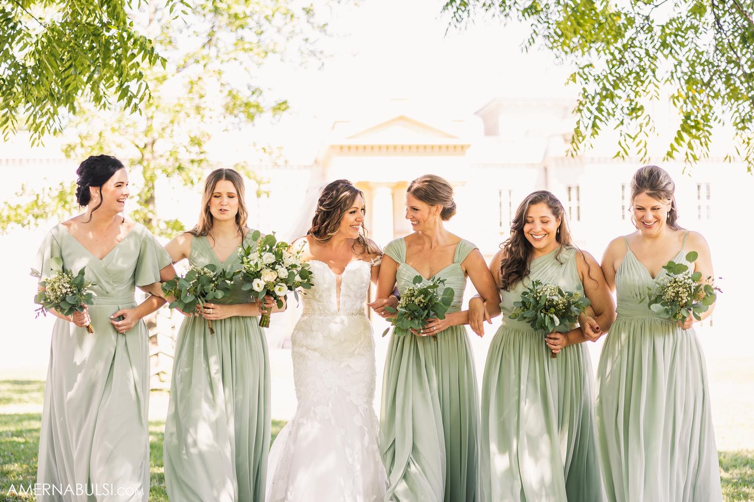 Bridesmaids in sage green dresses pose with bride holding white and green bouquets outdoors.