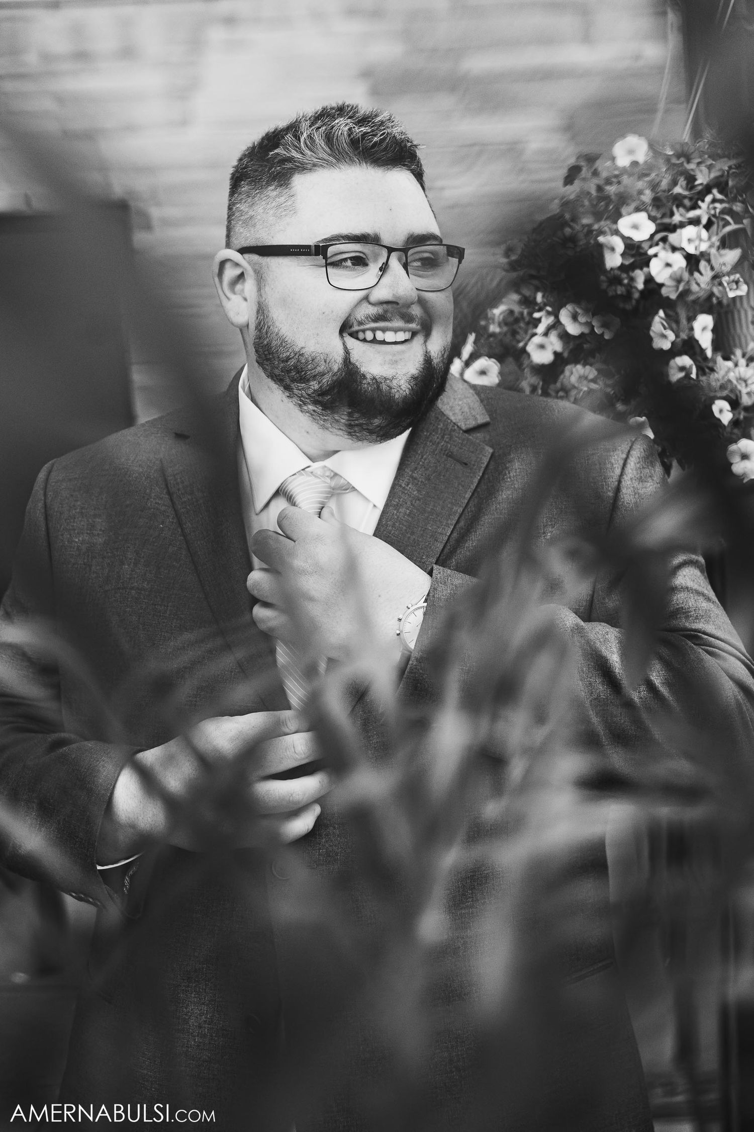 Black and white portrait of a bearded groom in glasses smiling while adjusting his tie.
